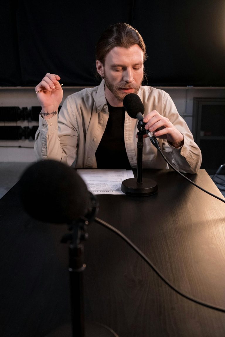 A bearded man recording a podcast indoors, engaging at a microphone stand with paper notes on a table.