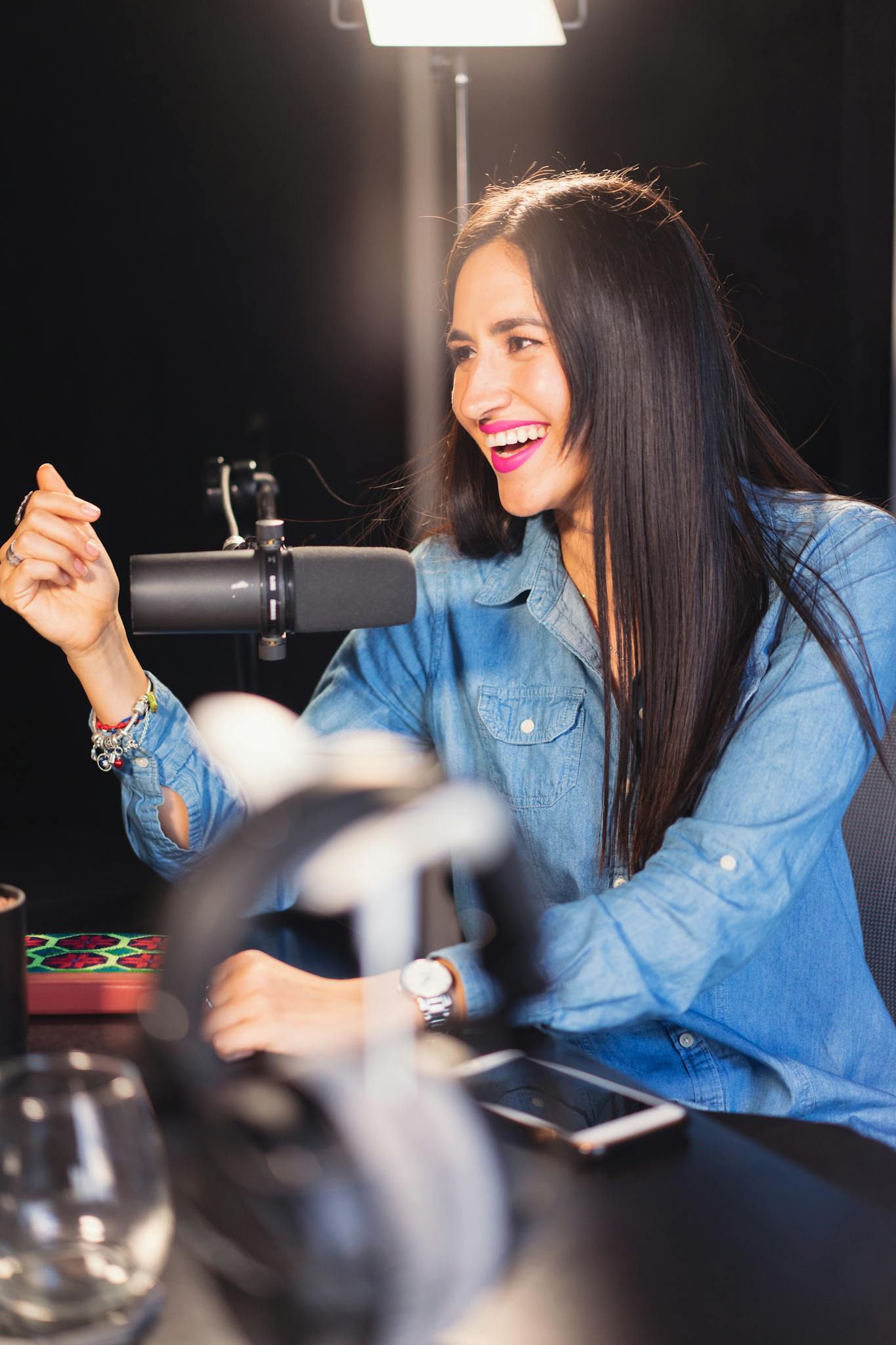 Smiling woman in denim recording a lively podcast in a professional studio setting.